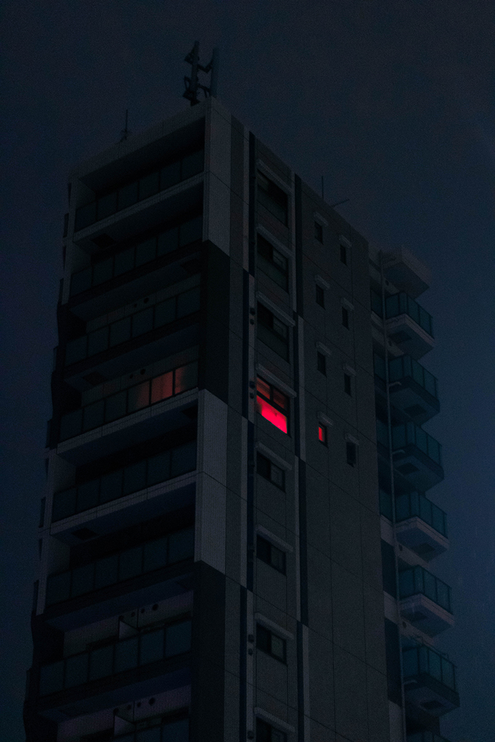 Dark foggy modern building with a single illuminated window in red and the dark cloudy sky behind it.